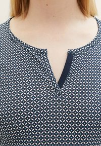 Haut marine avec un motif floral bleu et blanc. Présente un col en V et un tissu texturé. Manches courtes visibles.