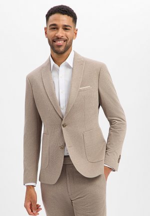 Man glimlachend, draagt een lichtbruine colbert en broek met een wit overhemd, staand met één hand in de zak tegen een witte achtergrond.
