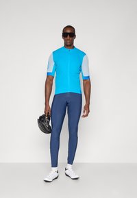 Maillot de cyclisme bleu à manches courtes, associé à un legging marine. Le modèle tient un casque noir. Des chaussures de cyclisme blanches complètent l'ensemble.