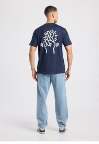 T-shirt en coton bleu marine avec un motif blanc représentant une main avec les doigts croisés dans le dos. Associé à un jean en denim bleu clair.