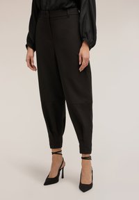 Pantalones negros a medida con cintura alta, piernas entalladas y textura suave, combinados con tacones altos negros de tiras.