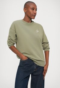 Sudadera verde oliva de tejido suave con cuello redondo, mangas enrolladas y un pequeño detalle del logo blanco en el pecho. Combinada con jeans azul oscuro.