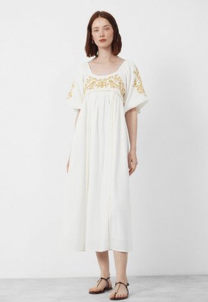 Vrouw met rood haar, gekleed in een witte midi-jurk met gele bloemembroidery en bruine platte sandalen, staand tegen een effen achtergrond.