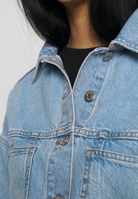 Närbild av en person som bär en ljusblå denimjacka över en svart skjorta, som visar kragen, knappar och bröstfickor.