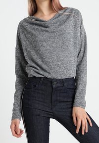 Maglietta grigia a maniche lunghe con scollo drappeggiato, abbinata a jeans in denim scuro con taglio aderente e dettagli a striscia laterale.