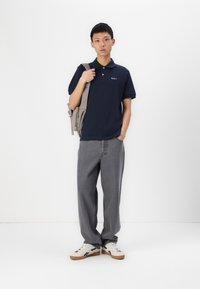 Jūras zila polo krekls ar īsu apkakli un logo, savienots ar brīviem pelēkiem džinsiem un baltām sporta kurpēm ar melniem akcentiem. Modelis nēsā pelēku somu.