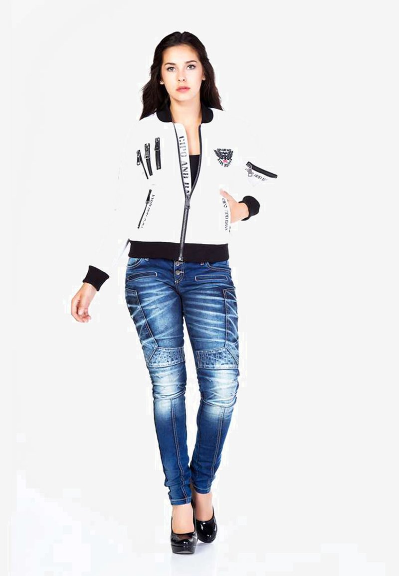 Veste bomber blanche avec des accents noirs et des détails graphiques, associée à un jean en denim bleu avec une texture déchirée et un design ajusté.