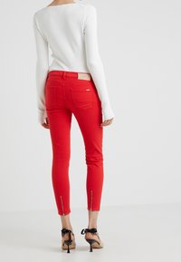 Persona que lleva pantalones rojos ajustados con bolsillos traseros y cremalleras en los tobillos, combinados con una blusa blanca de manga larga y tacones beige con cintas negras.