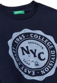 Námořnická modrá mikina se zaobleným potiskem s nápisem "NYC" v černé barvě, s opotřebovanými detaily; ribbed výstřih a viditelná zelená visačka značky.