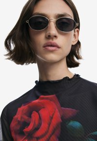 Schwarzes, durchscheinendes Oberteil mit einem großen roten Rosendruck, runde, dunkle Sonnenbrille mit goldenen Rahmen und einem Rüschendetail am Ausschnitt.