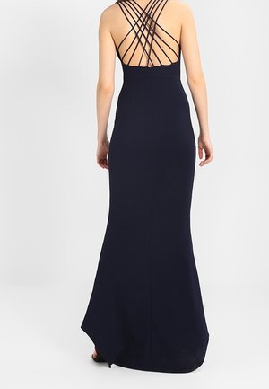 Femme portant une longue robe noire avec un dos ouvert à bretelles croisées, debout devant un fond clair uni.
