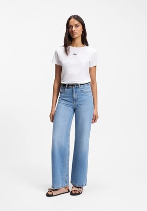 Donna in piedi che indossa una t-shirt bianca con scritta, jeans larghi a vita alta azzurri, cintura nera e sandali flat su sfondo bianco.