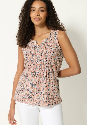 Kvinde med krøllet hår iført en ærmeløs blomstret bluse med knapper og hvide bukser, stående mod en ensfarvet baggrund.