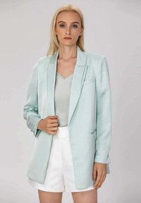 Blazer vert menthe avec revers crantés et deux poches avant, assorti à un haut gris clair et à un short blanc, texture lisse, coupe cintrée.