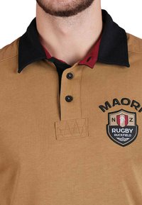 Polo sportif en rugby marron avec col noir, doublure intérieure rouge, trois boutons et un écusson Maori Rugby sur la poitrine. Tissu texturé.