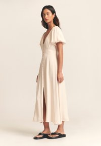 Lichtbeige jurk tot op de knie met diepe V-hals, pofmouwen en knoopsluiting aan de voorkant. Heeft een zijsplit en een gladde stoftextuur.