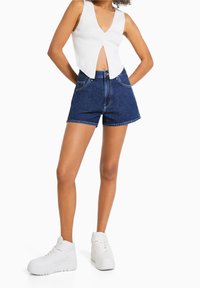 Tmavě modré džínové šortky s vysokým pasem, doplněné bílým žebrovaným tílkem s knoflíky a bílými vysokými teniskami.