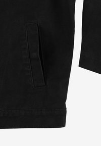 Czarna skórzana kurtka dżinsowa z boczną kieszenią, teksturowanym materiałem oraz wykończeniem przy mankietach i dolnym brzegu. Prosty, nowoczesny design.