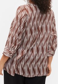 Donna che indossa una blusa a maniche lunghe marrone e bianca con i polsini arrotolati, vista da dietro, abbinata a pantaloni neri.