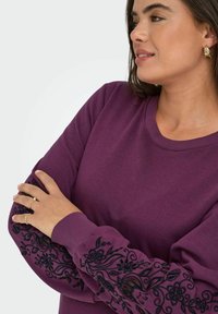 Sweat-shirt violet avec des manches longues ornées de broderies florales noires au niveau des poignets, affichant un col rond et une texture de tissu douce.