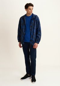 Giacca a vento blu camouflage con cappuccio, indossata sopra una felpa blu uniforme e pantaloni blu navy. Le scarpe sono sneakers nere con suole bianche.