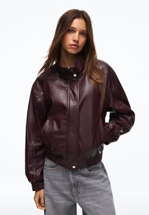 Junge Frau mit langen Haaren, die eine dunkelbraune Lederjacke und hellgraue Jeans trägt, steht vor einem weißen Hintergrund.