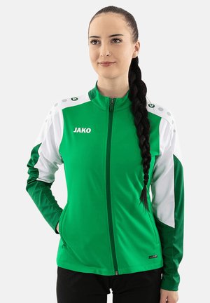 Donna con capelli lunghi intrecciati che indossa una giacca sportiva JAKO verde e bianca, in piedi con una mano in tasca, sfondo neutro.