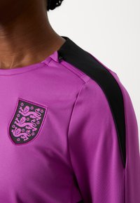 Lila sportpóló fekete válldíszítéssel és hímzett címerrel, amely oroszlánokat és virágmotívumokat ábrázol. Puha, sima anyagú textúra.