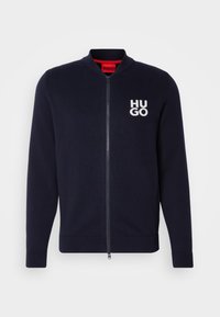 Marineblå cardigan med dragkedja, ribbade mudd och nederkant. Har en vit "HUGO"-logotyp på vänster bröst. Tillverkad av ett mjukt stickat tyg.