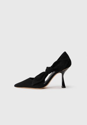 Scarpa nera in suede con tacco alto, punta affusolata e dettaglio a nodo sul lato. Presenta un tacco a stiletto lucido e un design con ritagli.