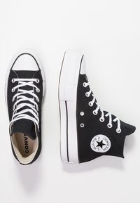 Czarne trampki typu high-top z białym noskiem i podeszwą, z białymi sznurówkami oraz okrągłą naszywką z logo po zewnętrznej stronie.