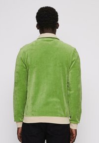 Zöld velúr dzseki puha textúrával, gallérral és bézs díszítéssel. Hosszú ujjakkal és enyhén ívelt derékvonallal rendelkezik.