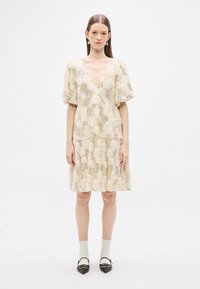 Ένα φόρεμα με κρεμ φλοράλ μοτίβο, ντεκολτέ σε σχήμα V, φουσκωτά μανίκια και φούστα με βολάν, κατασκευασμένο από υλικό με υφή που θυμίζει δαντέλα.