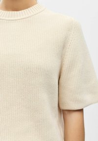 Pull en maille à manches courtes de couleur beige clair. Présente un col ras du cou côtelé et un tissu texturé avec une coupe ample.