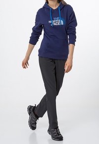 Granatowy sweatshirt z kapturem, z niebieskimi sznurkami i białym logo, zestawiony z czarnymi spodniami i czarnymi butami sportowymi. Prosty design, praktyczny krój.