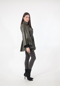 Olivgrüne, Kunstleder-Cape-Jacke mit weiten Ärmeln, an der Taille mit einem schwarzen Gürtel geschnürt. Kombiniert mit dunklen Skinny-Jeans und schwarzen Stiefeln.