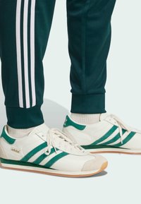 Weiße Sneakers mit grünen Wildlederakzenten und Streifen, ausgestattet mit einer strukturierten Zehenkappe und flacher Gummisohle, kombiniert mit grünen Sweatpants.