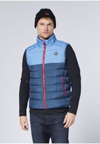 Blue and navy puffer vest with vertical stitching, red zipper, high collar, and logo badge. Worn over a black long-sleeve shirt.