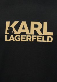 Svart stoff med beige "KARL LAGERFELD" tekst, med en silhuett av en person med solbriller og hestehale integrert i bokstaven K.