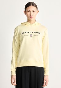 Žltá mikina s kapucňou z mäkkej tkaniny, s potlačou loga na prednej strane a klokanie kapsou, v kombinácii s čiernymi nohavicami.