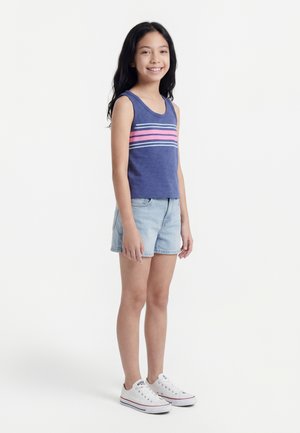 Κορίτσι που χαμογελά, φορώντας μπλε μπλούζα χωρίς μανίκια με ροζ και λευκές ρίγες, ανοιχτόχρωμα τζιν σορτς και λευκά αθλητικά παπούτσια, στέκεται μπροστά σε λευκό φόντο.