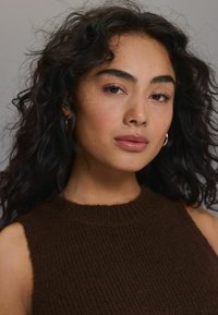 Braunes gestricktes ärmelloses Oberteil mit gerippter Textur, Rundhalsausschnitt und lockeren Locken, die das Gesicht einer Person mit leichtem Make-up umrahmen.