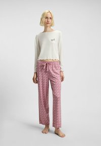 Witte lange mouwen top met een gerimpelde zoom en logo, gecombineerd met roze pyjamabroek met een herhalend patroon en gerimpelde manchetten.