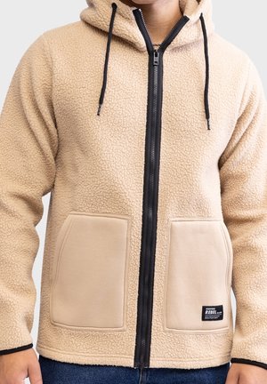 Chaqueta de forro polar sherpa beige con cremallera, capucha, cordones negros y dos grandes bolsillos delanteros, usada por una persona con las manos visibles.