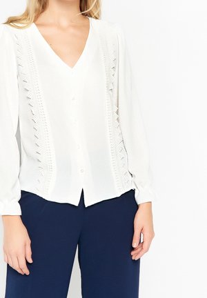 Kvinde iført en hvid langærmet bluse med blonde detaljer og marineblå bukser mod en ensfarvet hvid baggrund.