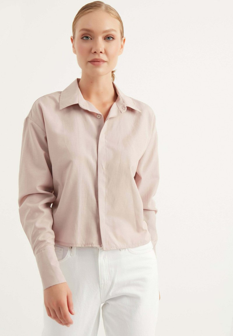 Camicia a maniche lunghe rosa chiaro con colletto a bottoni, modello corto e texture liscia, abbinata a pantaloni bianchi.