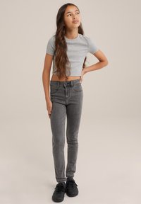 Grå kortärmad t-shirt, högmidjade mörkgrå skinny jeans med kontrasterande sömmar, bärs med svarta sneakers. Stående pose mot en neutral bakgrund.