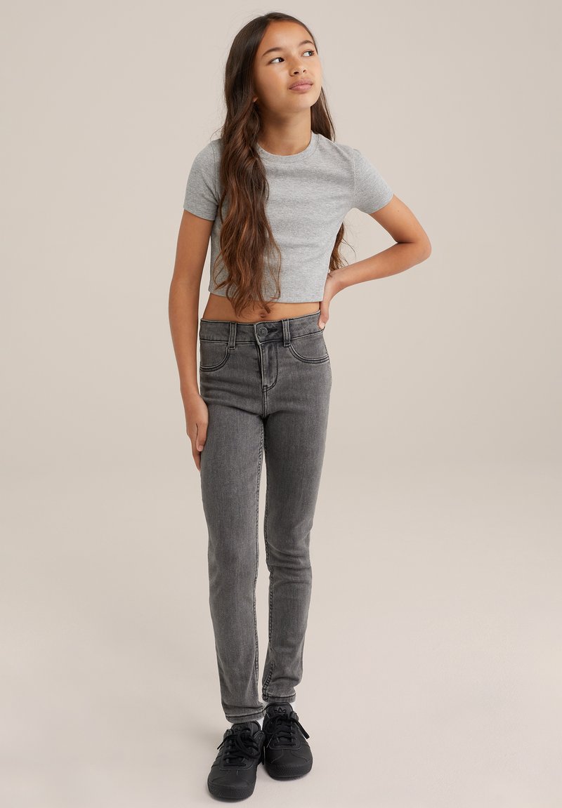 Grå kortärmad t-shirt, högmidjade mörkgrå skinny jeans med kontrasterande sömmar, bärs med svarta sneakers. Stående pose mot en neutral bakgrund.
