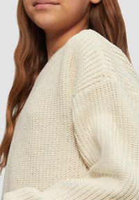 Sweter w kolorze kremowym z dzianiny, z teksturowanym wzorem, okrągłym dekoltem i opadającymi ramionami. Miękki materiał widoczny w luźnym kroju.
