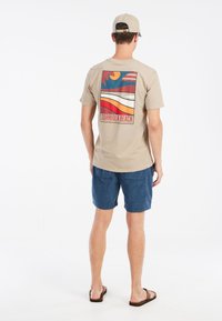 camiseta beige de manga corta con un diseño gráfico colorido en la parte de atrás que incluye patrones ondulados; combinada con pantalones cortos azules y sandalias.